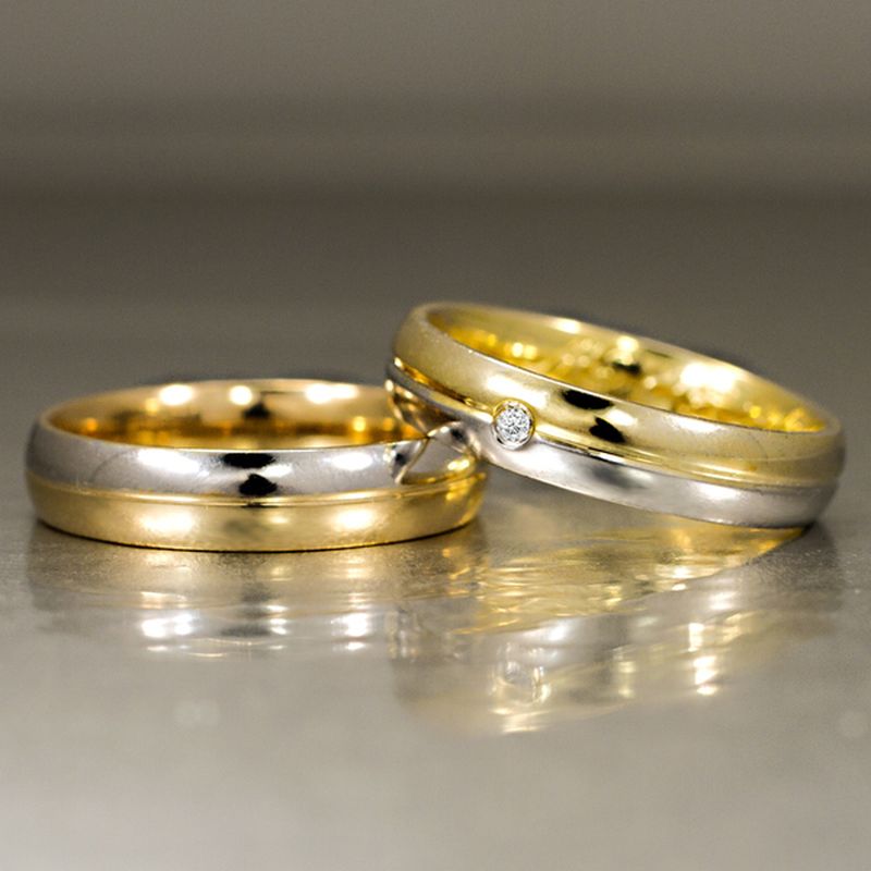 Duas alianças, uma com a ponta sobre a outra, feitas em ouro branco e amarelo, com uma pedrinha branca.