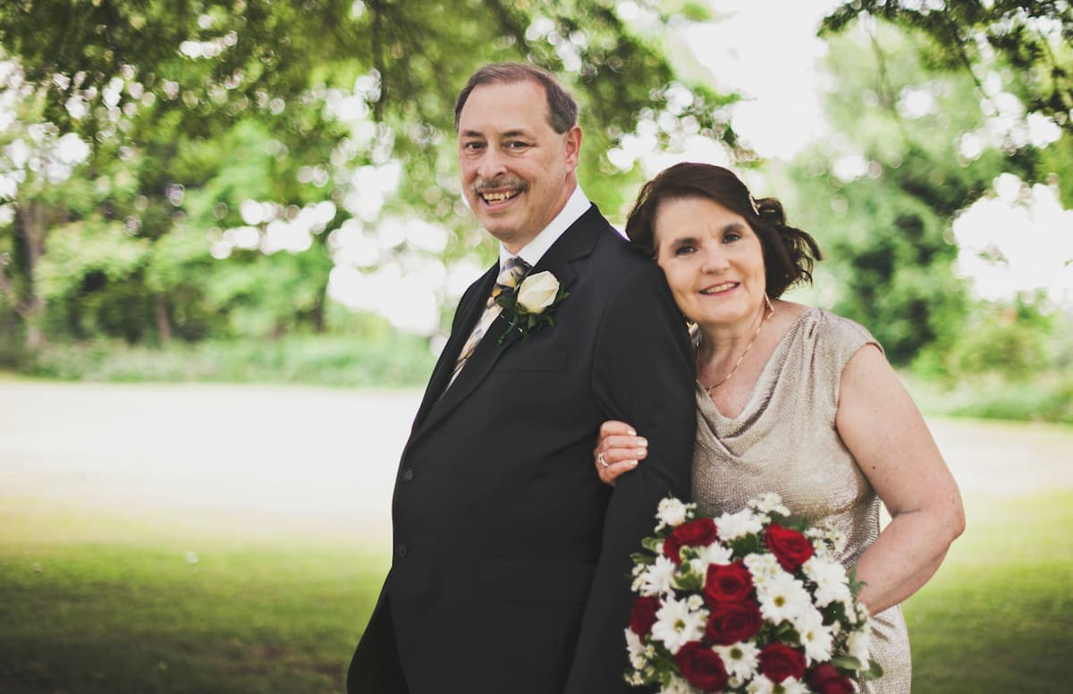 Imagem de um casal de pessoas mais velhas segurando um buquê de flores com roupa social