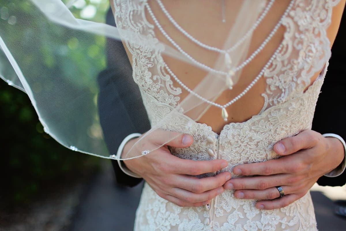 Imagem das costas de um vestido de noiva sendo abraçada pelo noivo