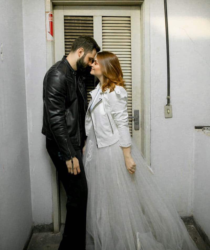Casal em look de ensaio de casamento, ela com vestido branco e longo, de casamento, e uma jaqueta de couro branca; ele com roupas pretas e jaqueta de couro preta