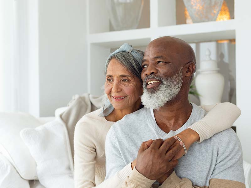 Front view of senior diverse couple sitting on a white couch in beach house. Authentic Senior Retired Life Concept