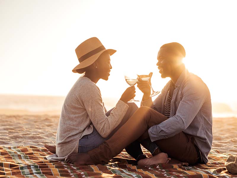 Romantic young African couple toasting each other with wine while sitting together on a sandy beach at dusk