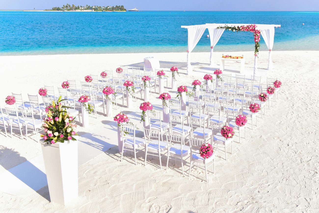recepção de casamento na areia. Foto mostra areia branca com cadeiras brancas organizadas em fikeiras e decoradas com flores rosas. Mar azul ao fundo