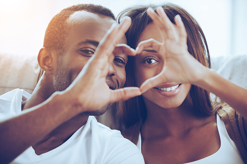 Love is in the air! Beautiful young African couple sitting close to each other and looking through a heart shape made with their fingers
