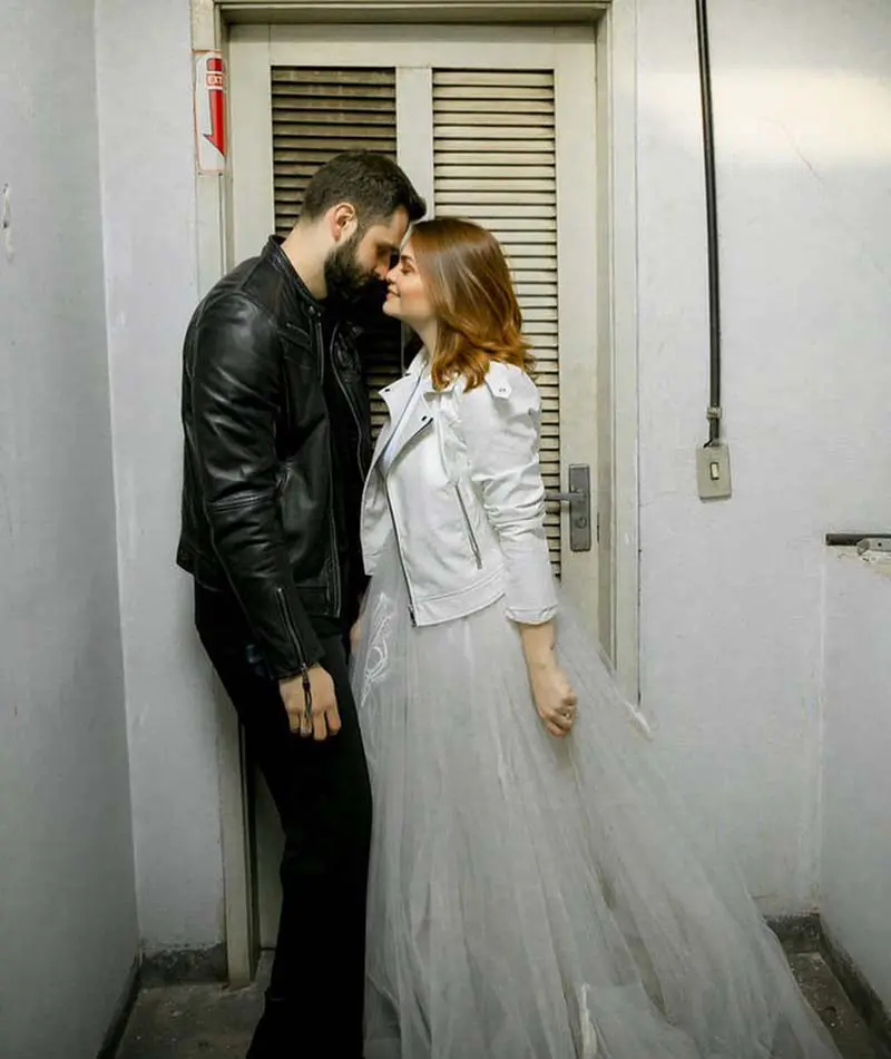 Casal em look de ensaio de casamento, ela com vestido branco e longo, de casamento, e uma jaqueta de couro branca; ele com roupas pretas e jaqueta de couro preta